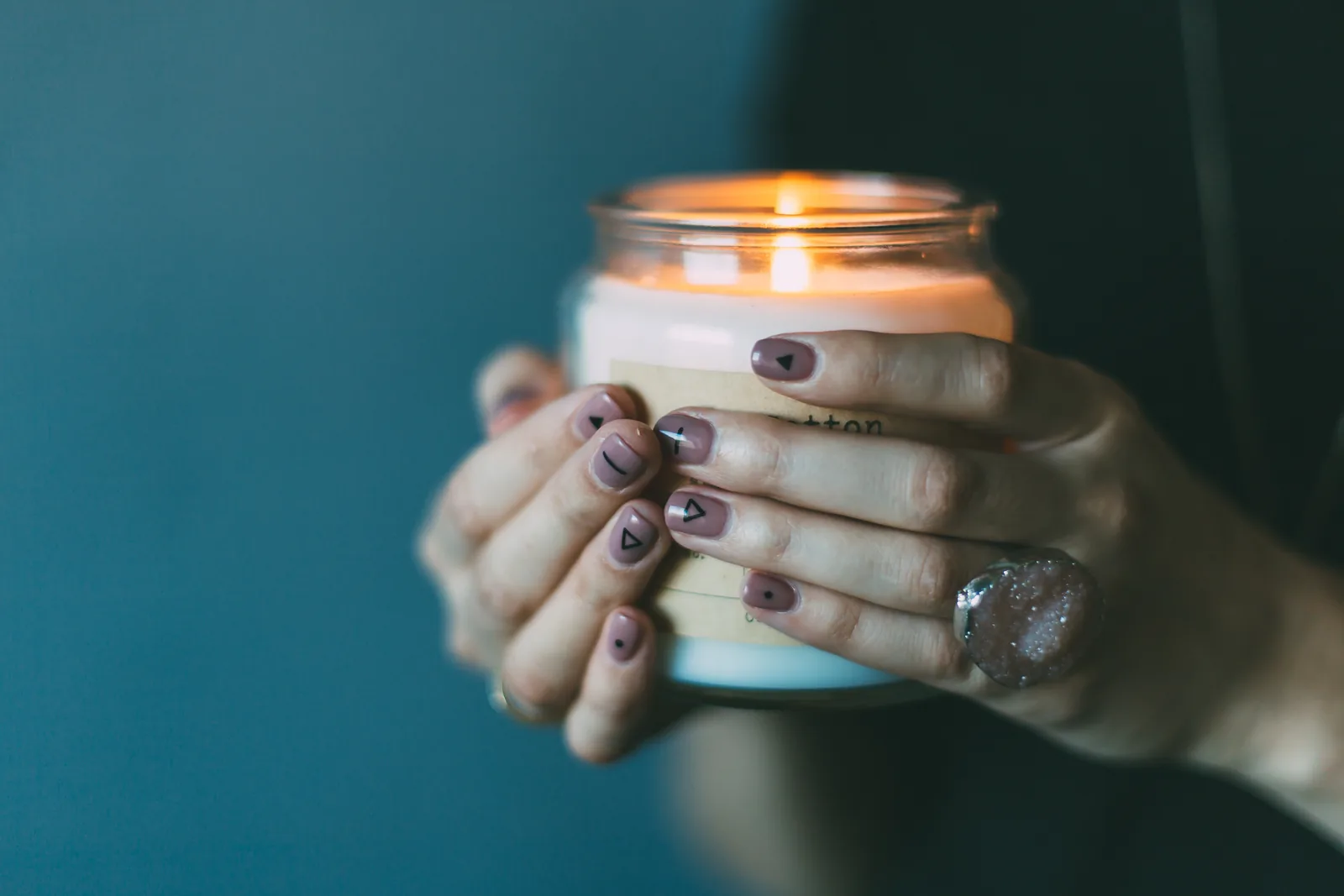 Hands with dark polished nails holding a lit candle in a jar